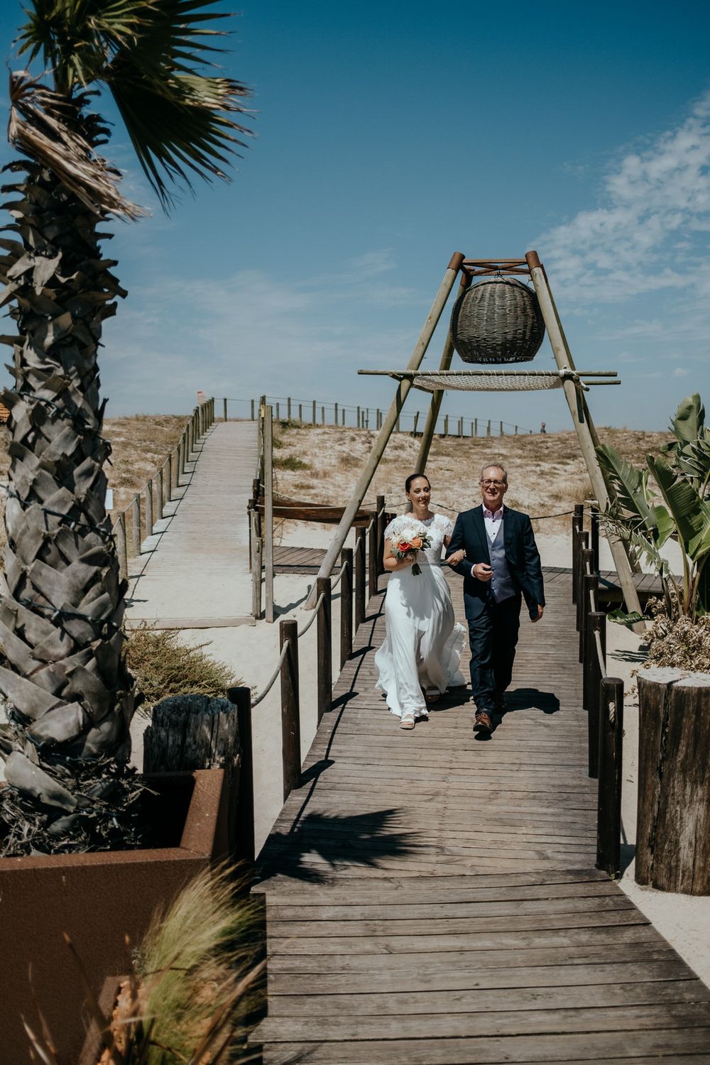 Bride and father walk down wooden boardwalk path at outdoor beach wedding ceremony under blue sky.