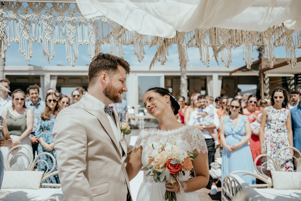 Happy couple exchanging vows at an outdoor wedding ceremony, bride holding a bouquet, guests celebrating in the background.