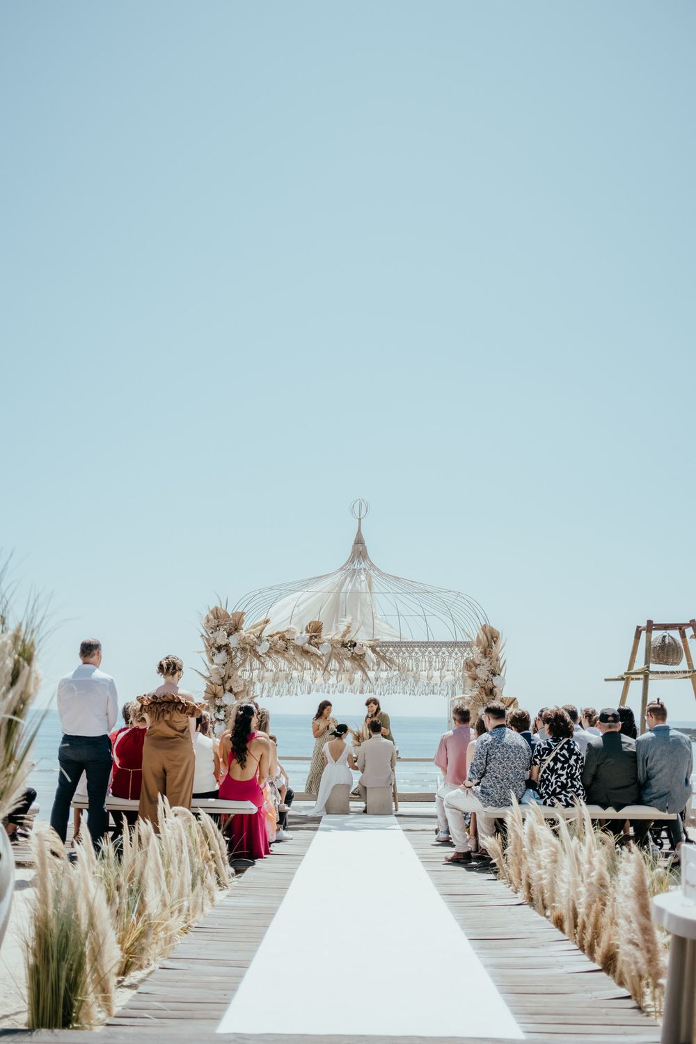 Outdoor beach wedding ceremony with boho macrame arch, white aisle runner, and pampas grass decor under a clear blue sky.