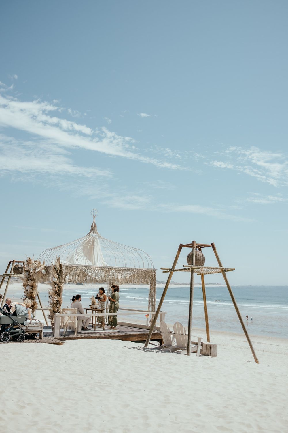 Bohemian beach wedding ceremony setup with wicker canopy, wooden structures, and guests on white sand under a blue sky.
