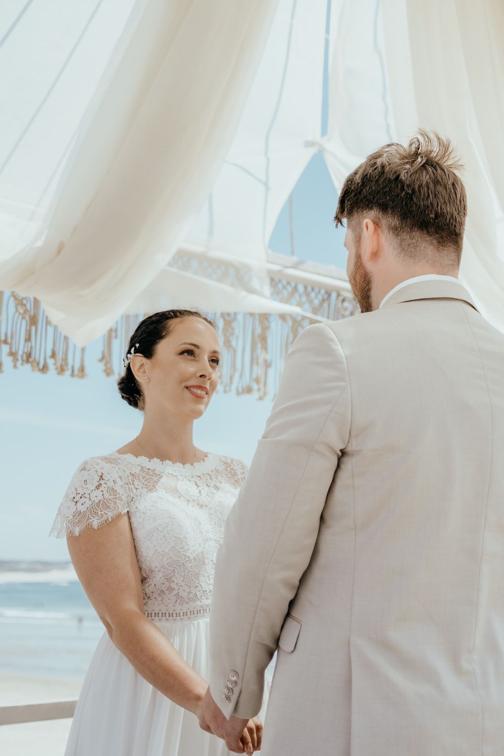Bride and groom holding hands at a beach wedding ceremony under white draping, exchanging vows by the ocean.