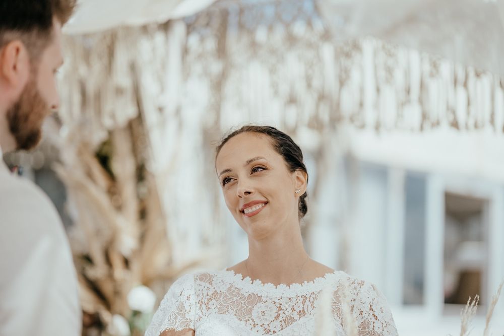 Bride in lace wedding dress smiling joyfully at groom during outdoor wedding ceremony.