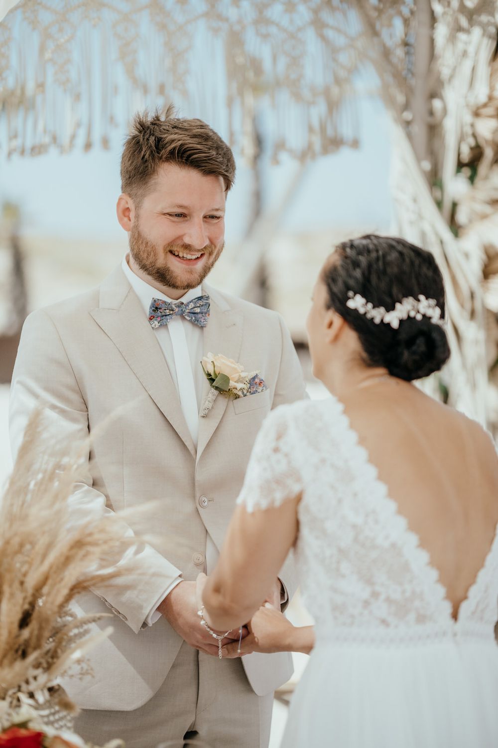 Smiling groom in beige suit with bow tie holds hands with bride in lace dress during outdoor beach wedding ceremony.