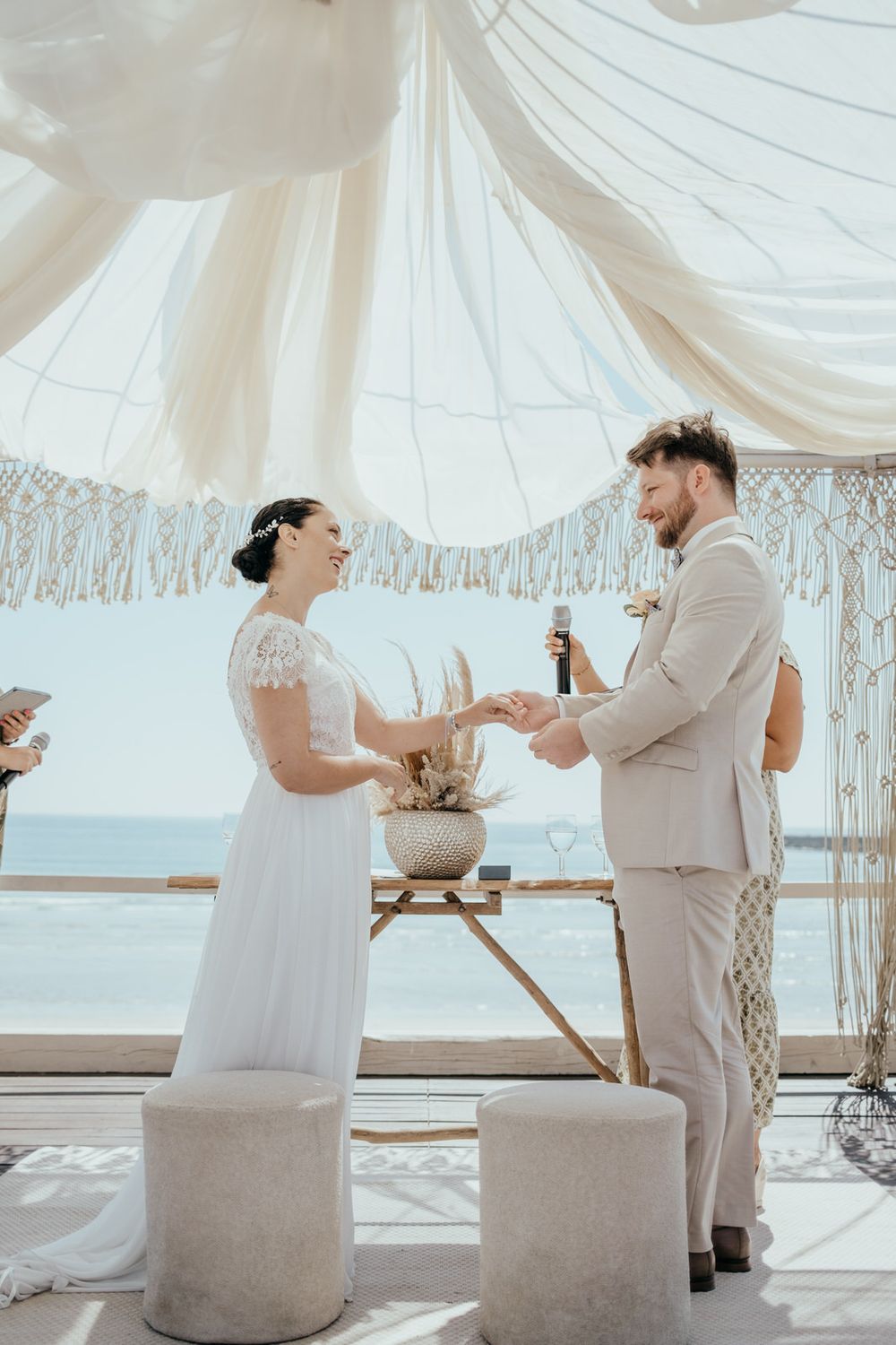 Bride and groom exchange rings at a boho beach wedding ceremony under white draped fabric with ocean views.