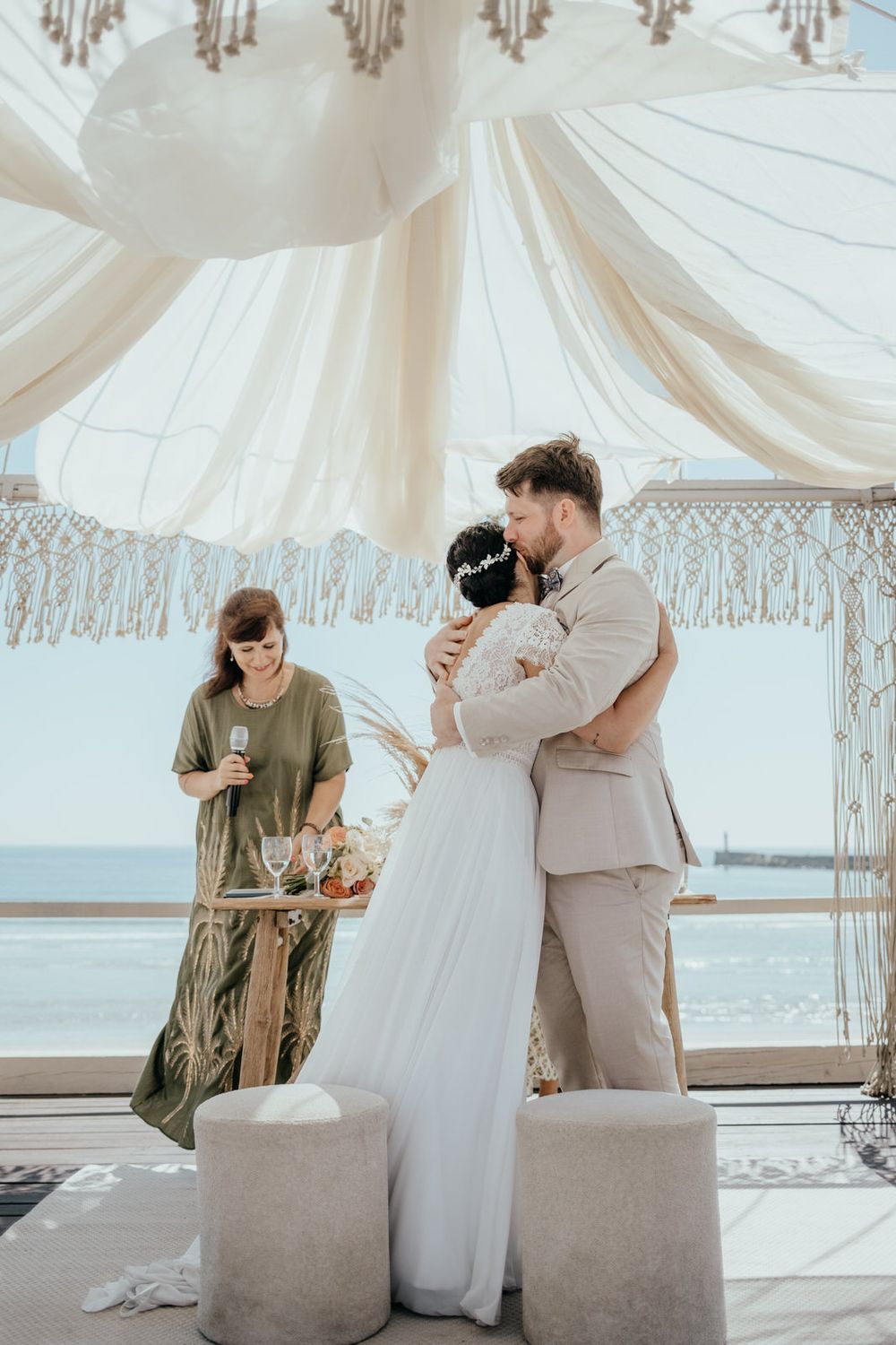 Bride and groom share first kiss at beachfront wedding ceremony under white macrame arch with officiant nearby.