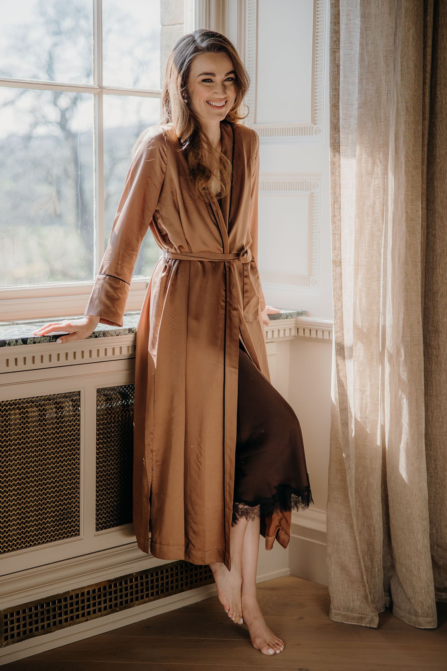 Woman smiling in a satin bronze robe with black lace trim, standing barefoot by a sunlit window.