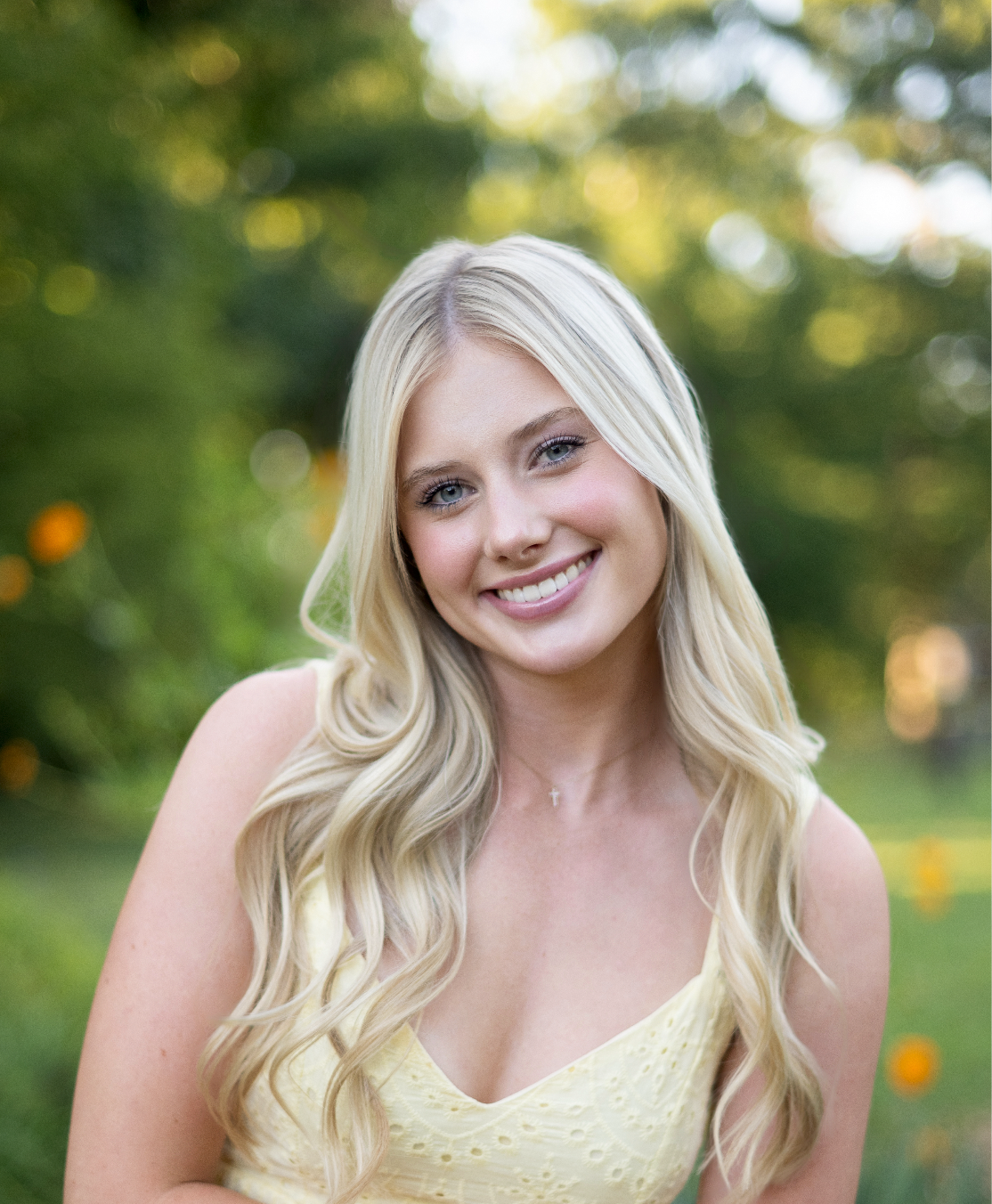 Young blonde woman smiling outdoors in yellow dress with bokeh green and orange floral background.