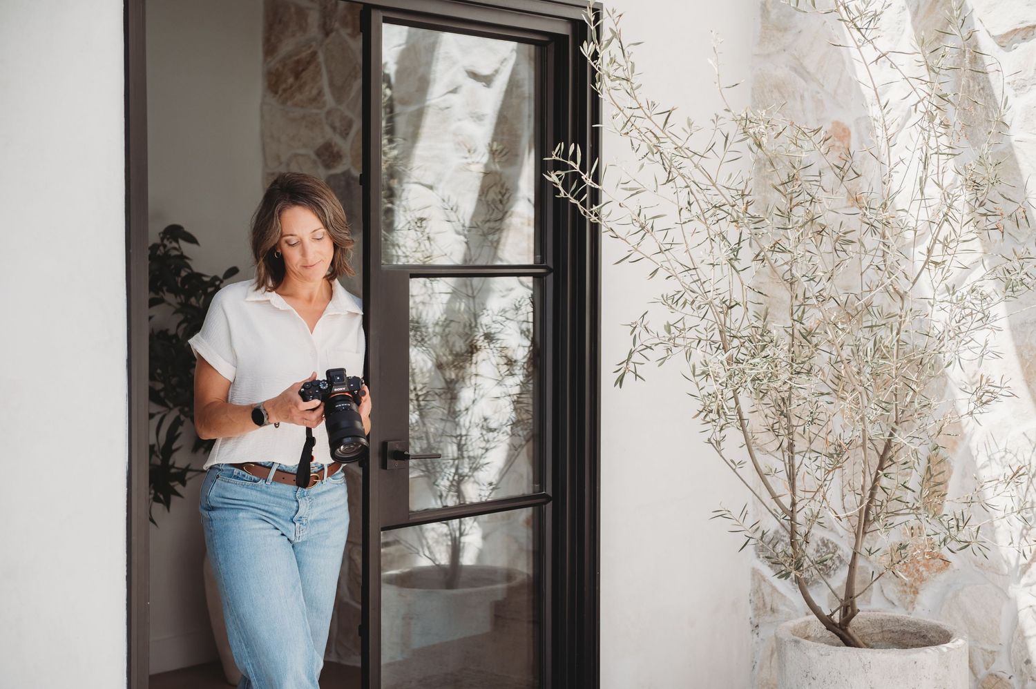 Woman in white shirt and jeans holding a camera, standing in a black-framed doorway with a plant nearby.