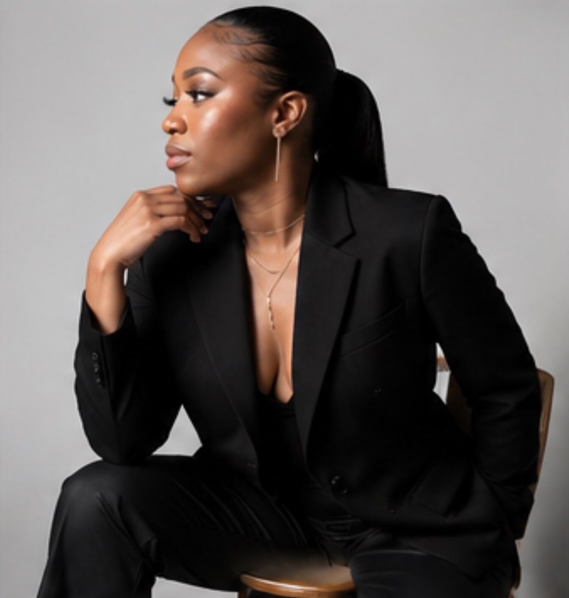 A woman in a black blazer suit sits elegantly, wearing gold earrings and necklace, posing against a gray background.