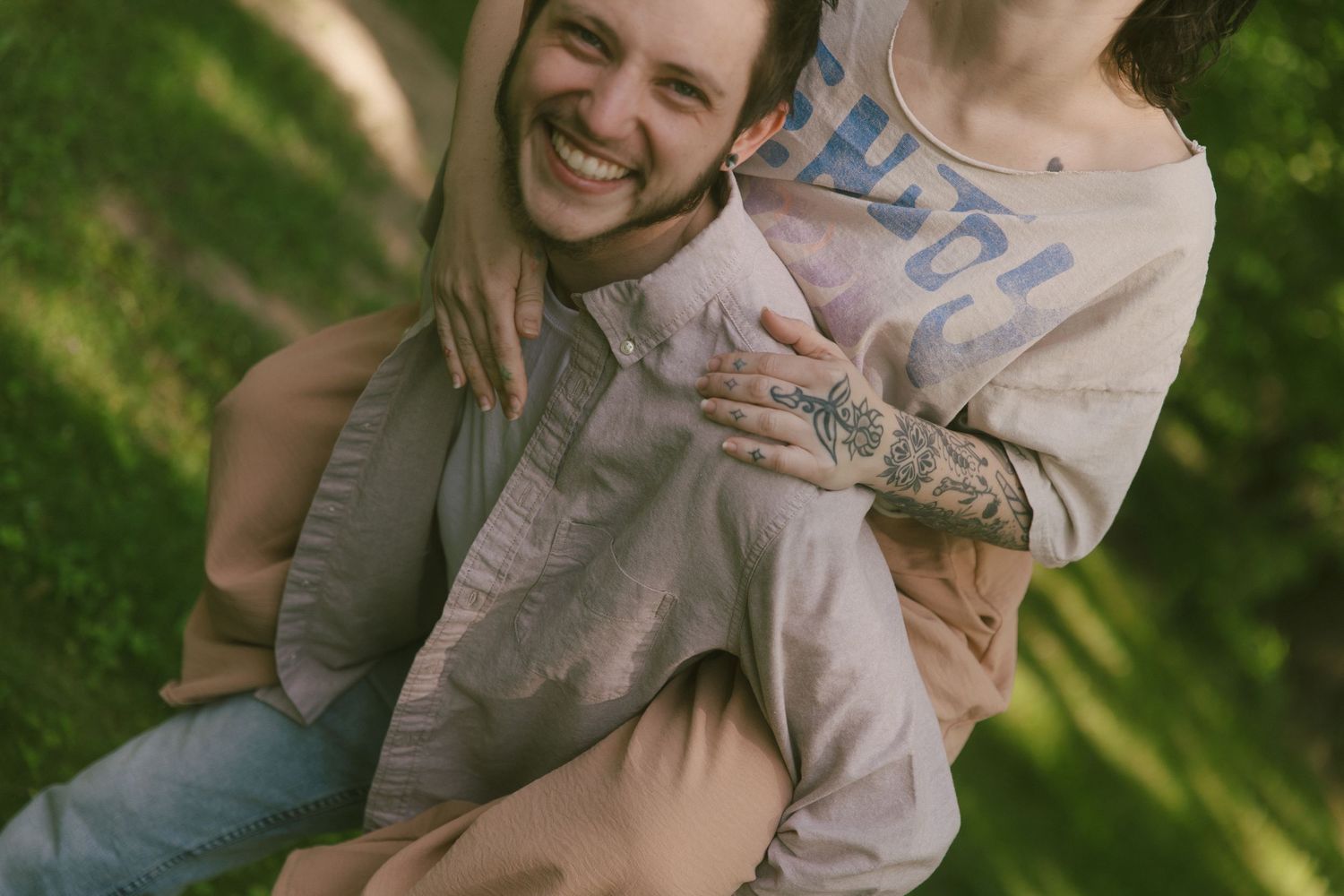 A laughing man with tattoos carries a woman on his back in a sunny green park setting.