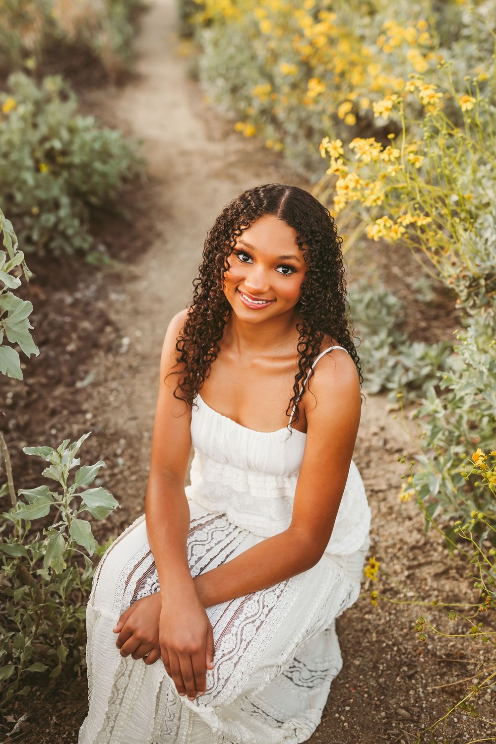 Smiling young woman with curly hair in white dress seated on a trail among yellow blooming flowers.