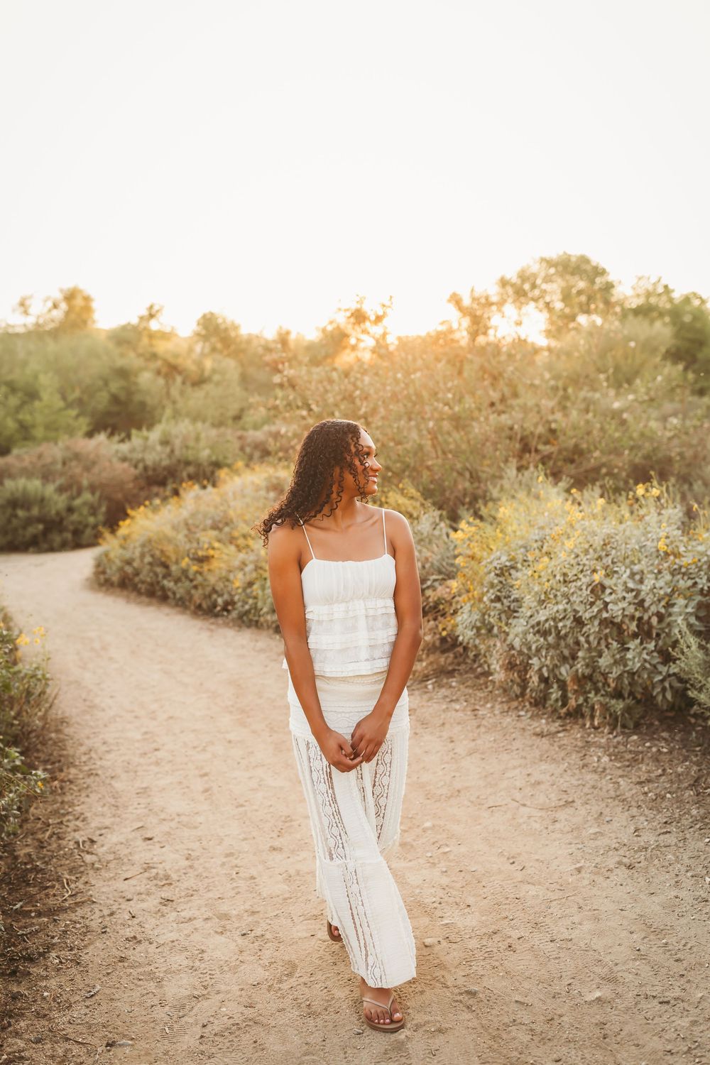 A young woman in a white lace maxi dress walks a sandy path surrounded by golden-hour lit greenery.