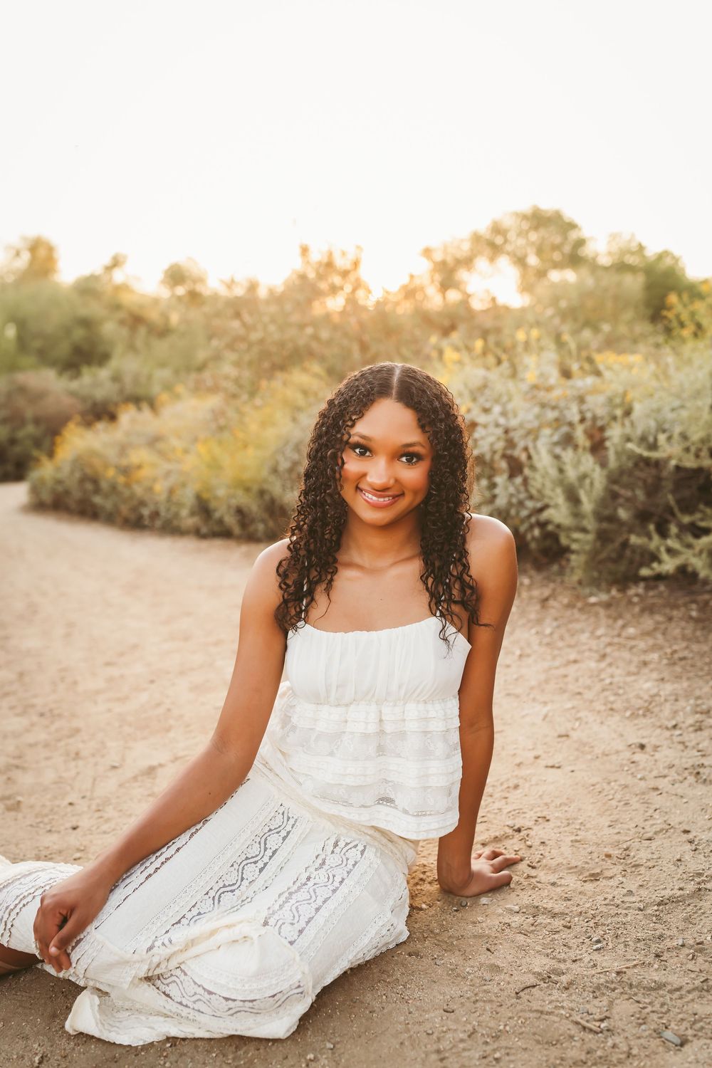 Young woman in white dress sitting on sandy path surrounded by golden hour greenery, smiling outdoors.