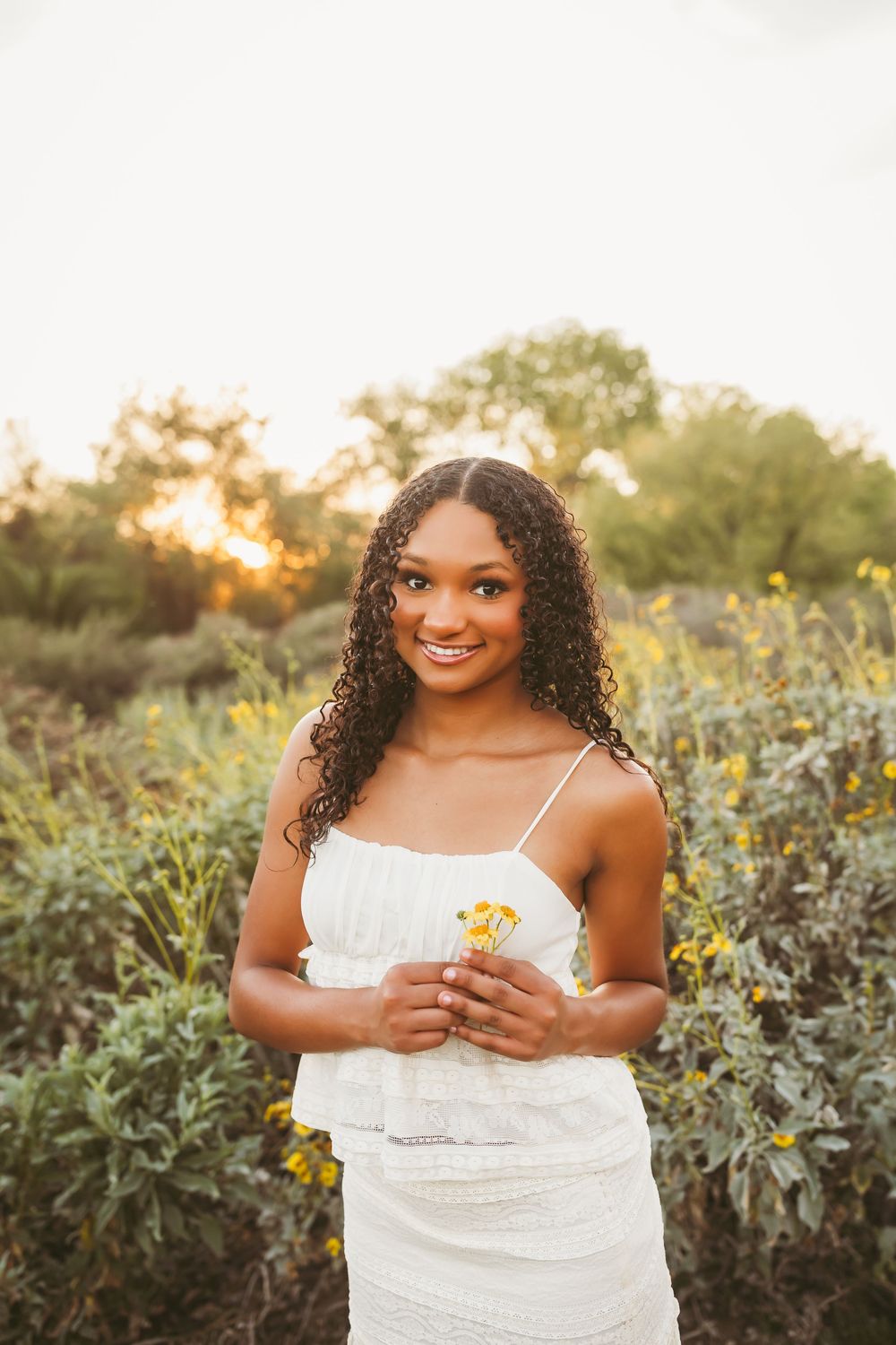 Young woman in white outfit holding yellow wildflowers while standing in a sunlit garden at golden hour.