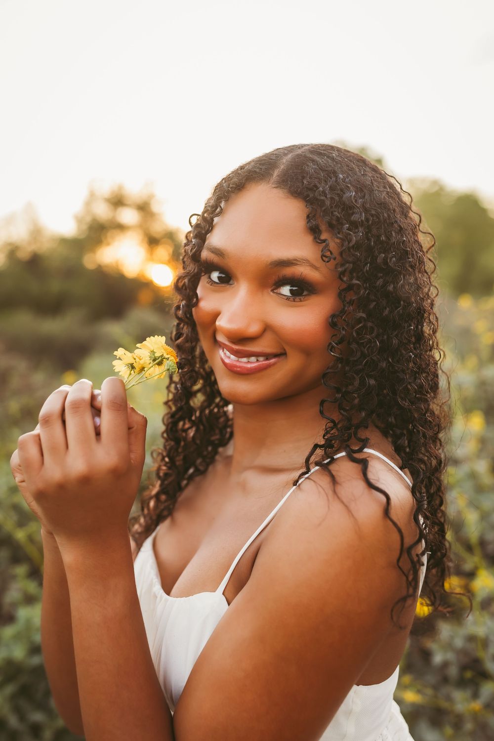 Young woman with curly hair holding yellow flowers, smiling outdoors in a white dress during golden hour.