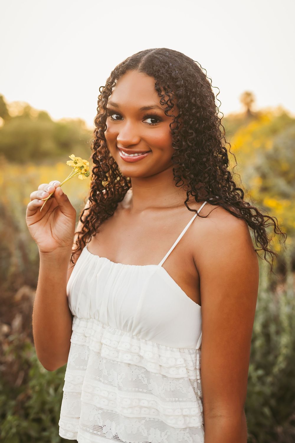 Young Black woman with curly hair in white dress holding yellow wildflower, smiling outdoors at golden hour.