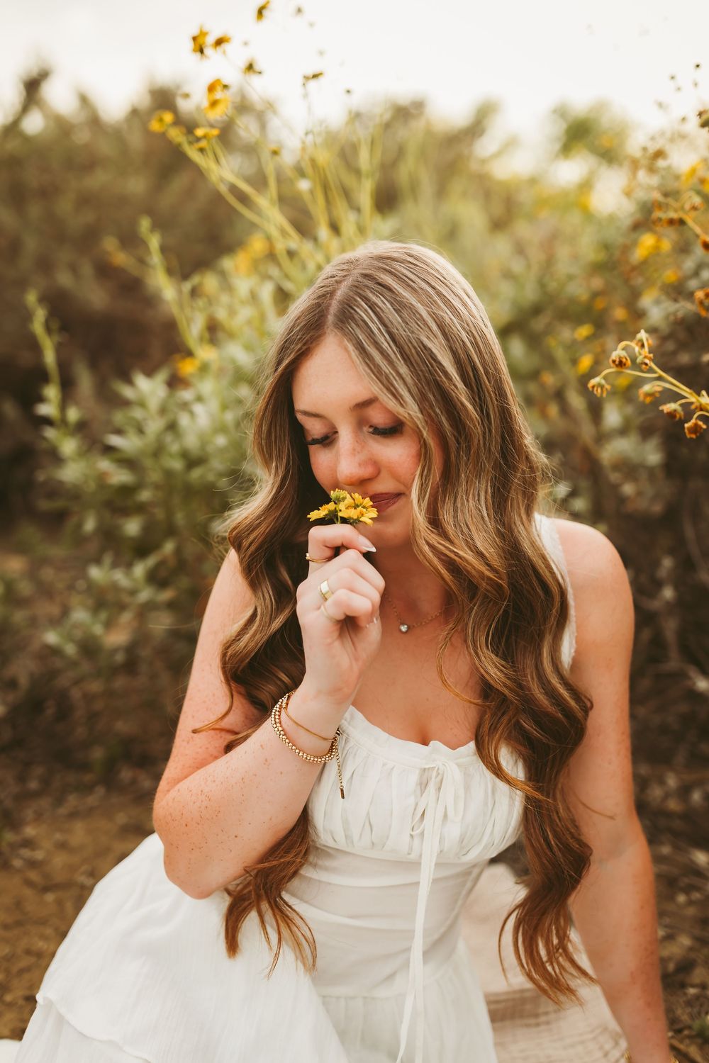 Young woman in white dress smelling yellow wildflowers outdoors, surrounded by lush greenery.