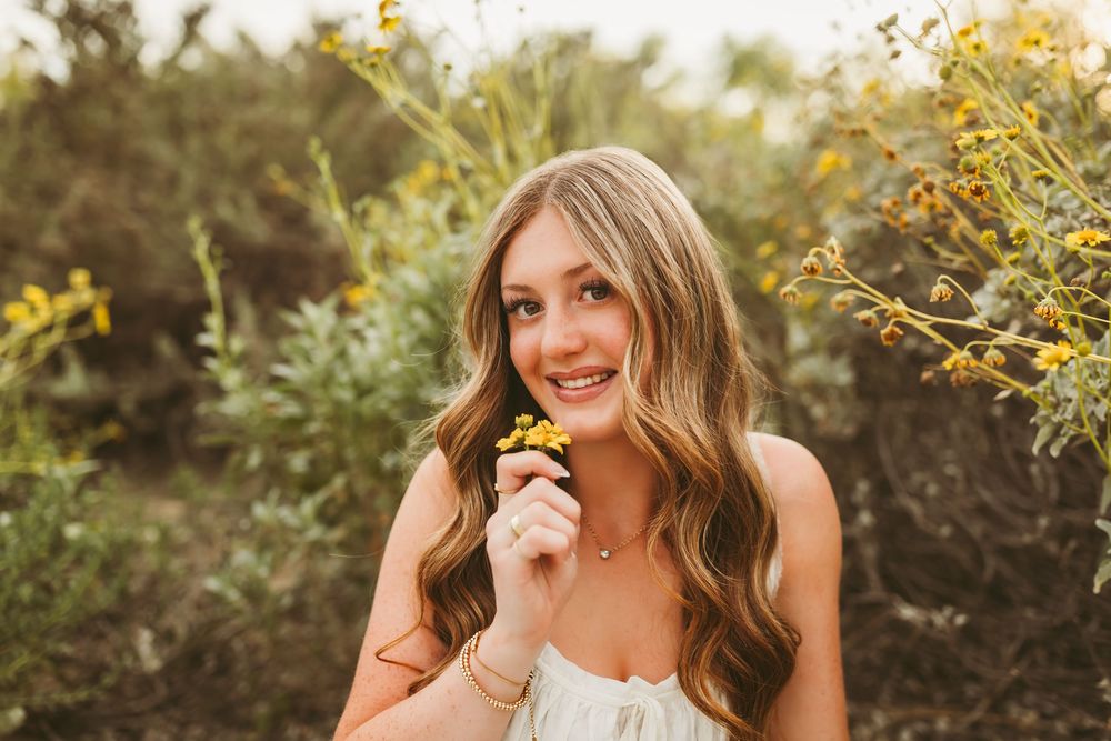 Young woman with long wavy hair smiling outdoors, holding yellow wildflowers among lush green bushes in warm sunlight.