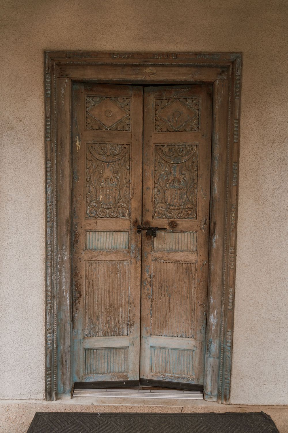 Antique carved wooden double door with ornate detailing and metal latch set in a rustic stucco wall.