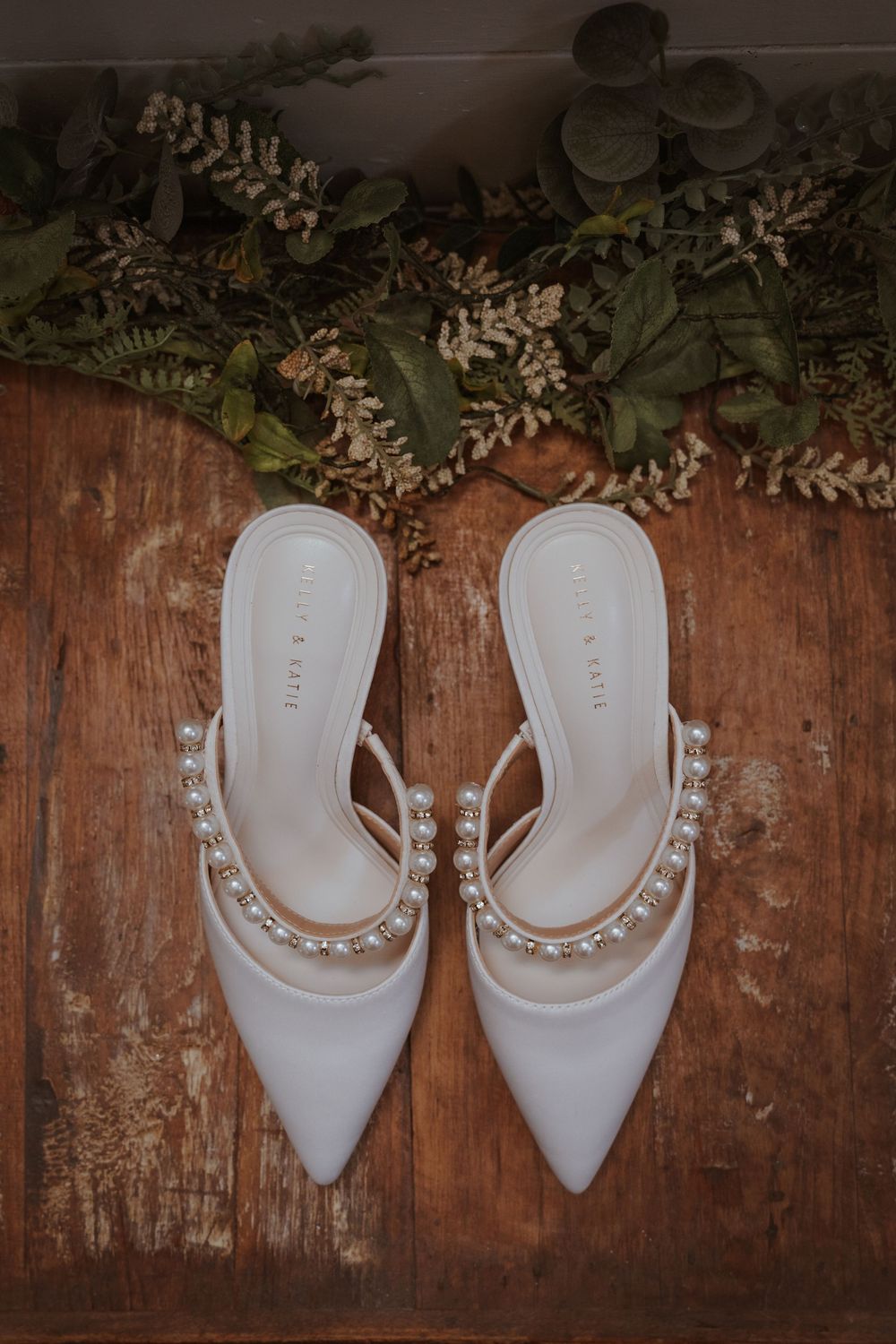 White pearl-embellished bridal heels on rustic wood with greenery.