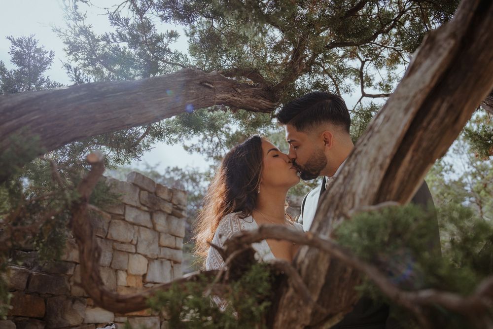 Couple sharing a romantic kiss among lush trees and stone ruins in an outdoor garden setting.