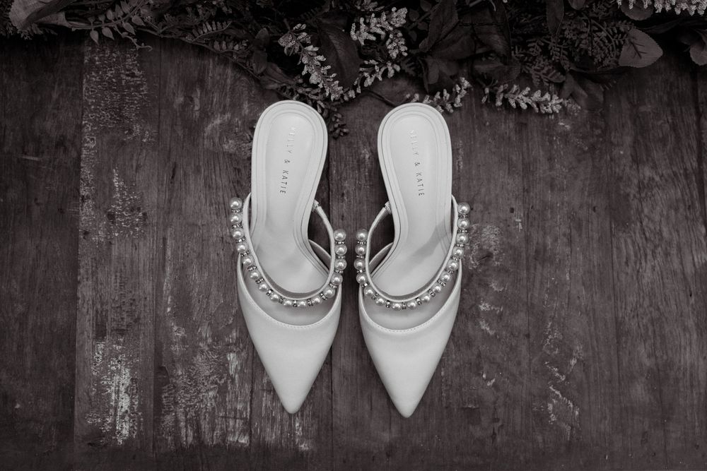 Elegant white bridal heels with pearl embellishments on rustic dark wood floor for wedding day.