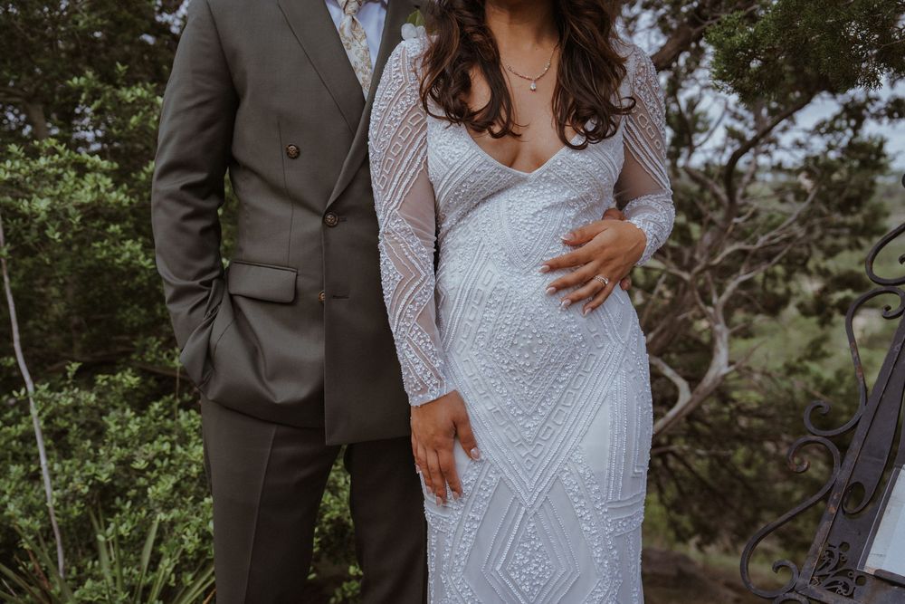 Elegant bride in long-sleeve lace wedding dress stands with groom in gray suit outdoors among green trees.