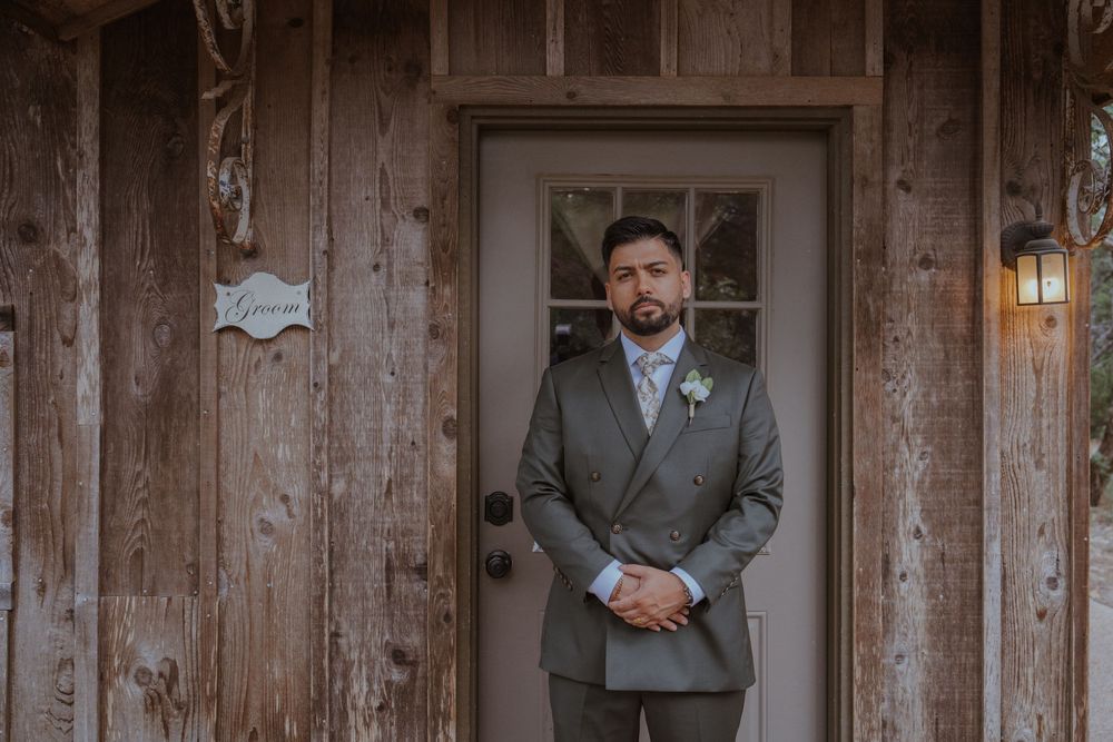 Groom in gray suit stands before rustic wooden door with Groom sign at outdoor wedding venue.