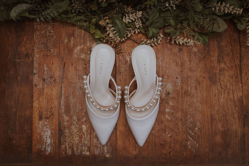 White pearl-embellished pointed-toe bridal mules on rustic wood floor with greenery.