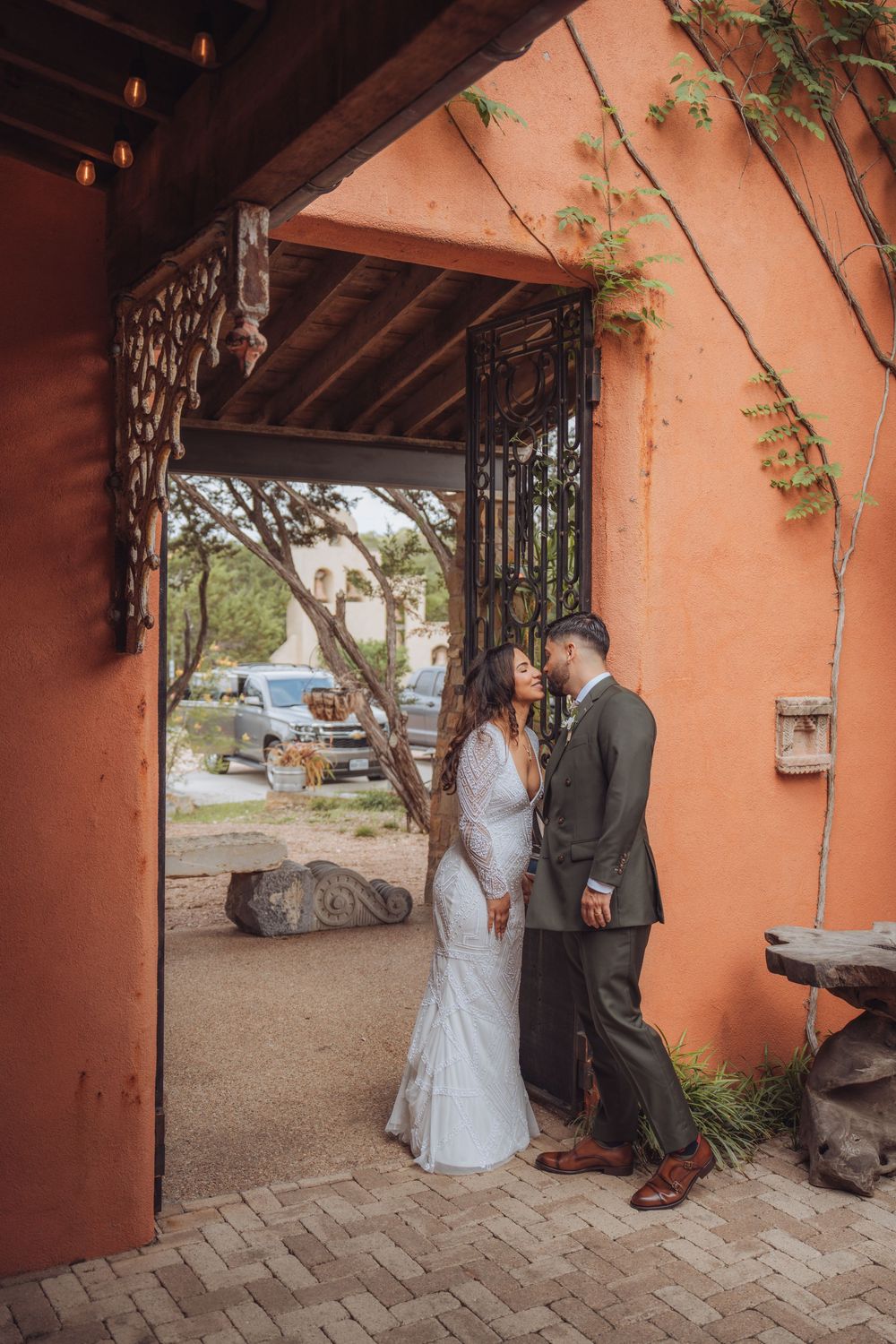 A bride and groom share an intimate moment under a rustic adobe archway with ornate iron gates.