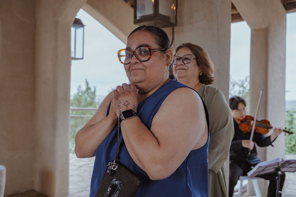 A woman in a blue sleeveless dress clasps her hands to her chest emotionally at an outdoor wedding event.