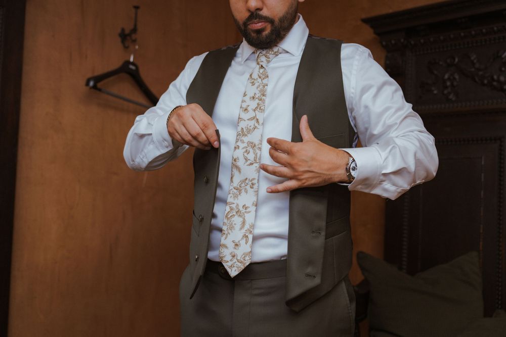 Man in gray vest and floral tie adjusting his outfit, wearing a watch, standing near wooden wall.