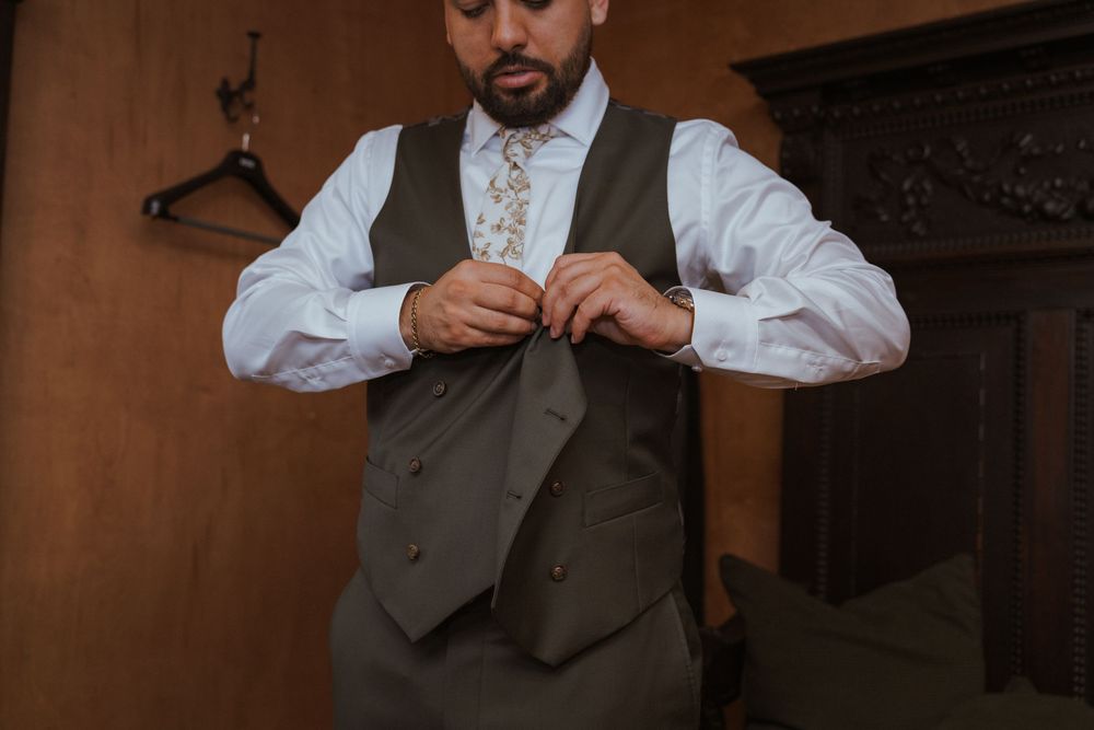 A groom in a gray vest and floral tie buttons his vest while getting ready for his wedding ceremony.