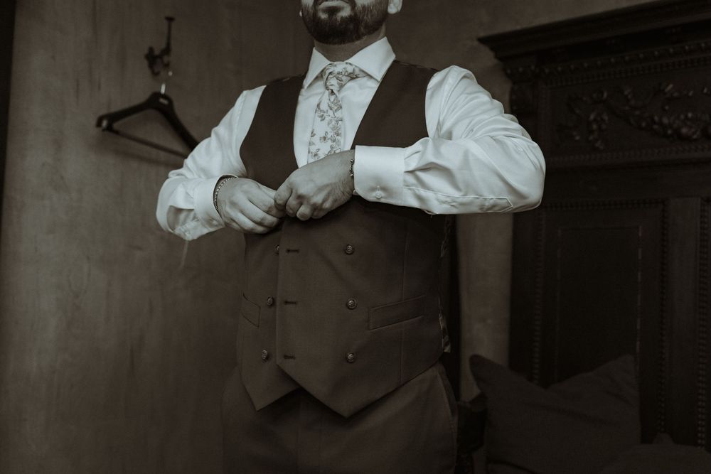 Groom buttoning his white dress shirt and floral tie under a brown vest during wedding preparations.