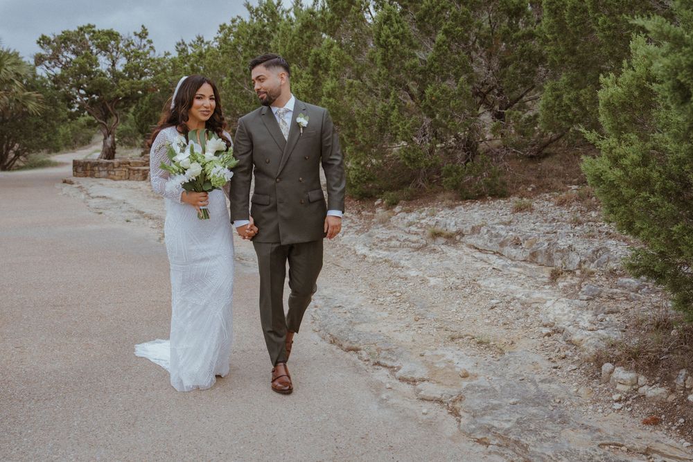 A bride and groom walk hand in hand along a scenic path surrounded by lush green trees on their wedding day.