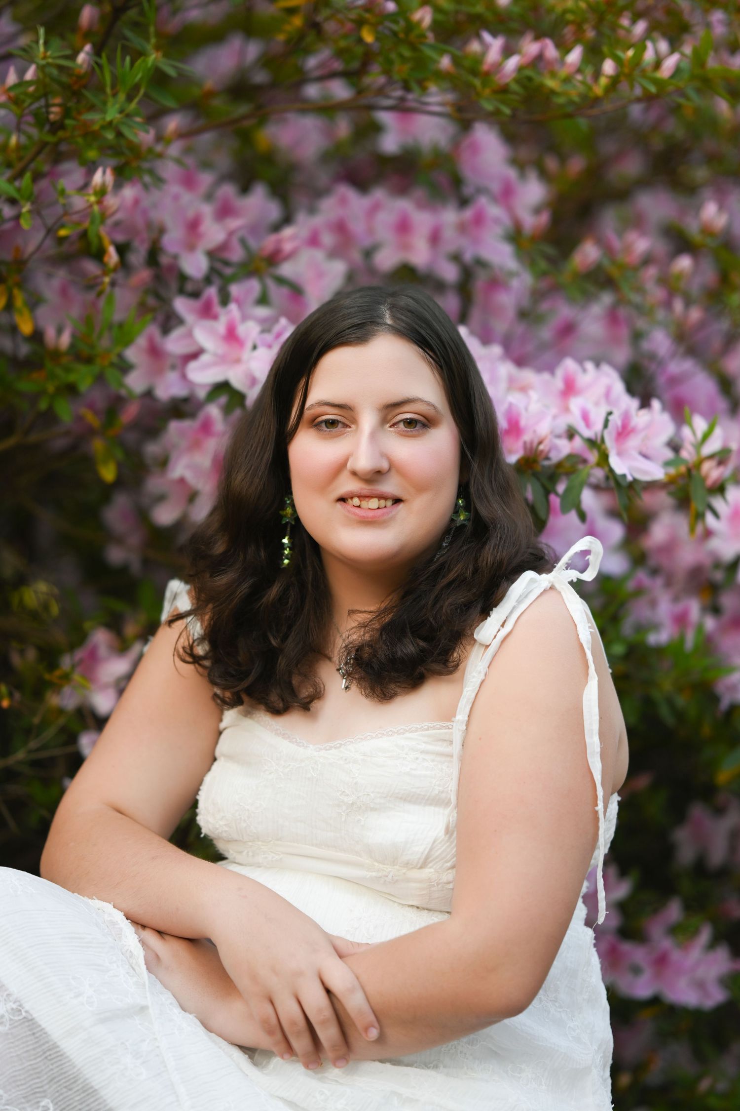 Woman in white dress smiling among pink azalea blooms, wearing green earrings in an outdoor garden setting.