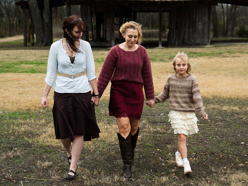 Three women and a girl walk hand-in-hand outdoors near a rustic barn on a grassy field.
