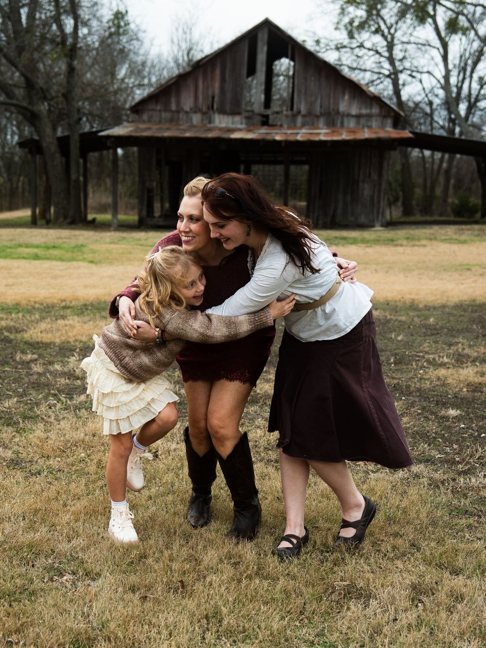 Three females share a warm group hug outdoors near a rustic barn on a cloudy day.