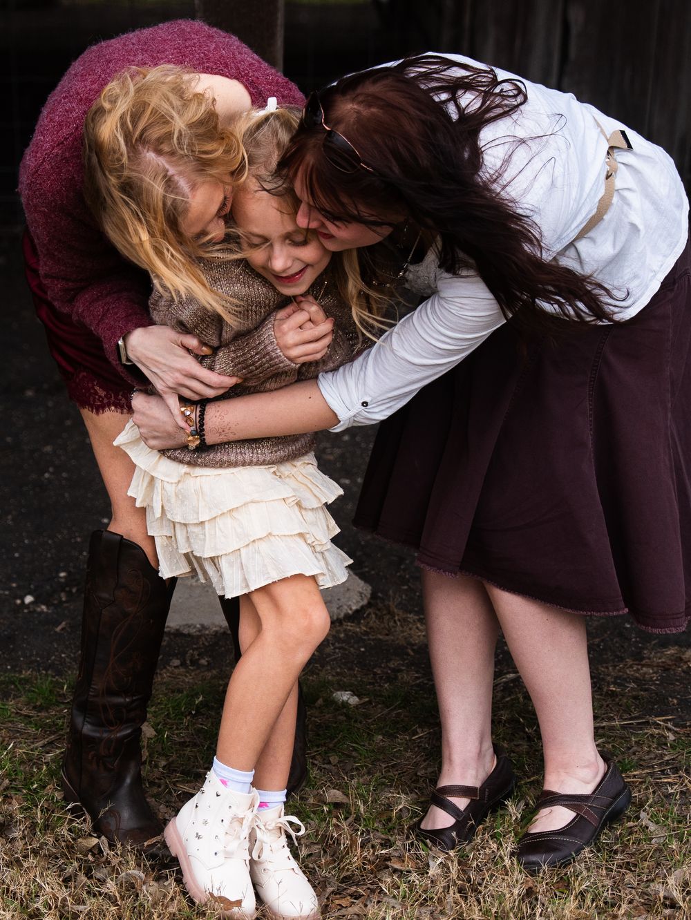 Two women hugging a young girl outdoors, sharing a warm embrace in casual clothing.