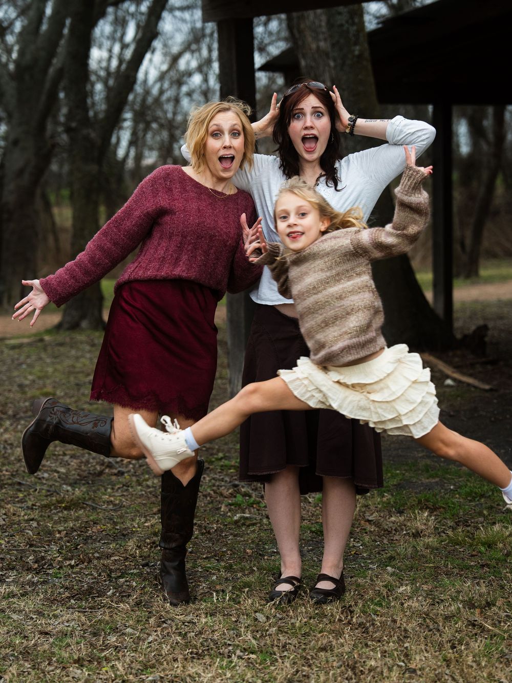 Two women and a young girl strike fun, surprised poses outdoors among trees in coordinated fall outfits.