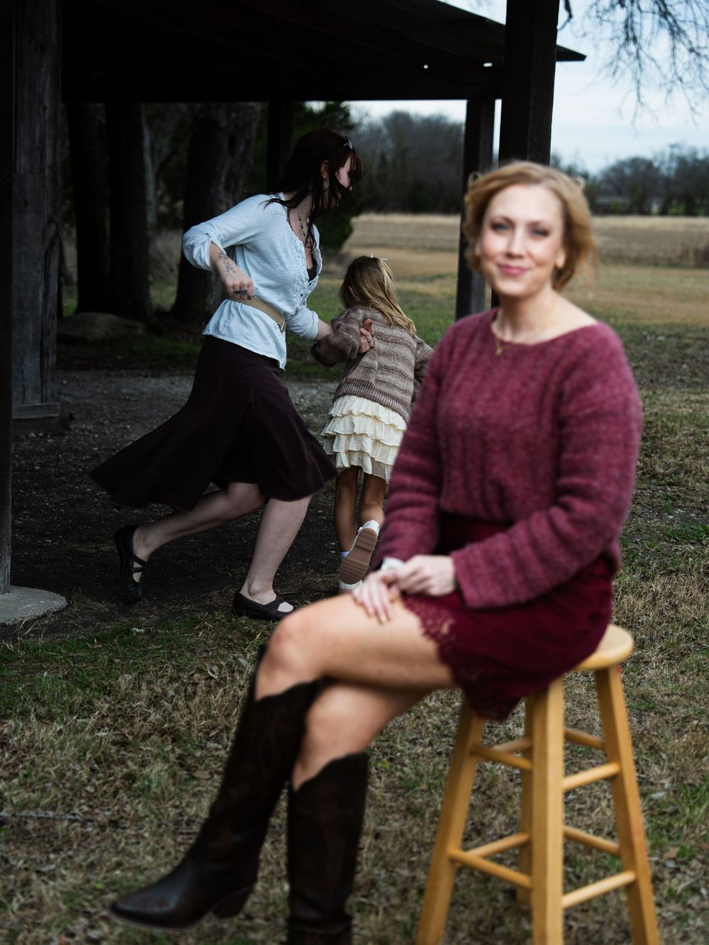 Woman in burgundy sweater dress and boots sits on stool outdoors while two girls play behind her.