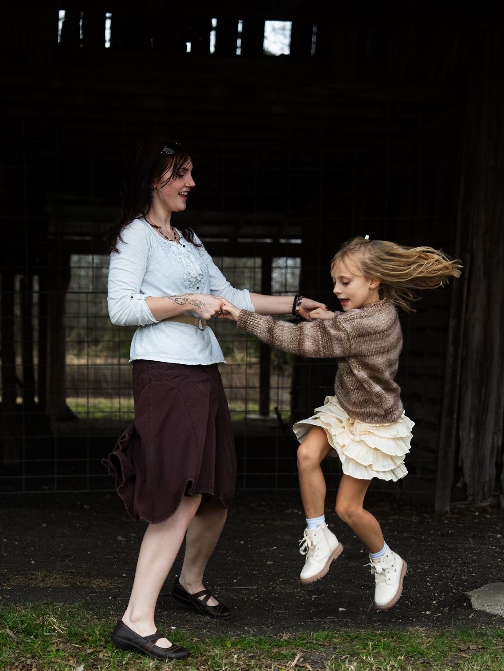 A woman and young girl joyfully dance together outdoors, with the girl leaping mid-air while holding hands.