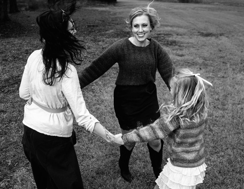 Three women holding hands in a circle outdoors, laughing and spinning together in a black and white photo.