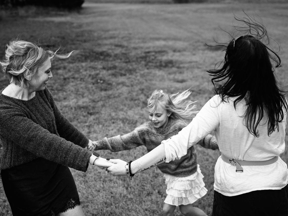 Black and white photo of two women and a young girl holding hands, spinning together outdoors on a grassy lawn.