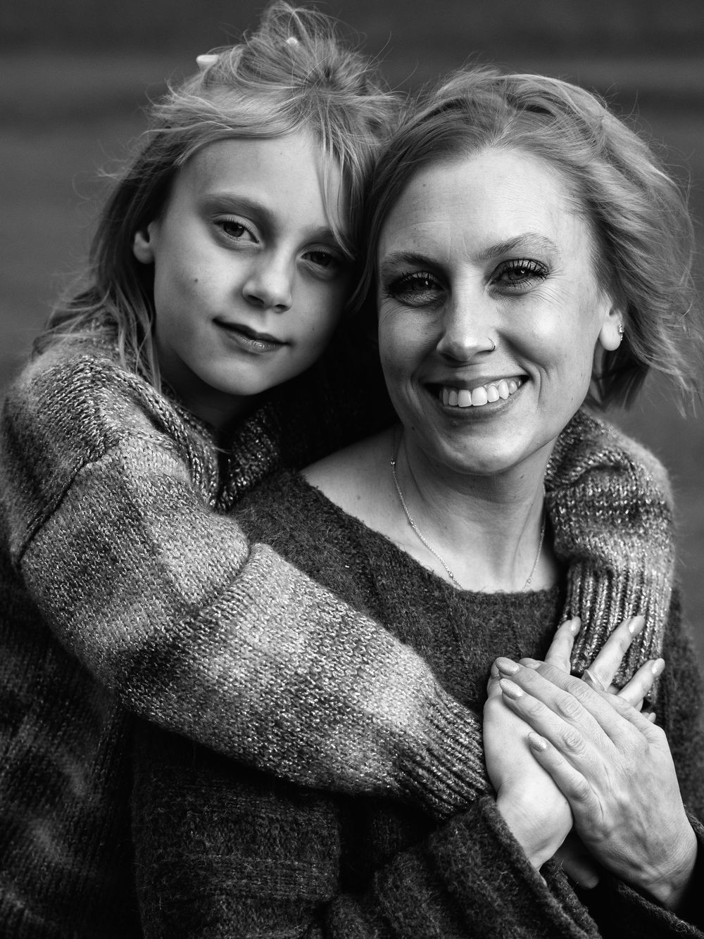 Black and white portrait of a smiling woman and young girl hugging warmly, both wearing cozy knit sweaters.