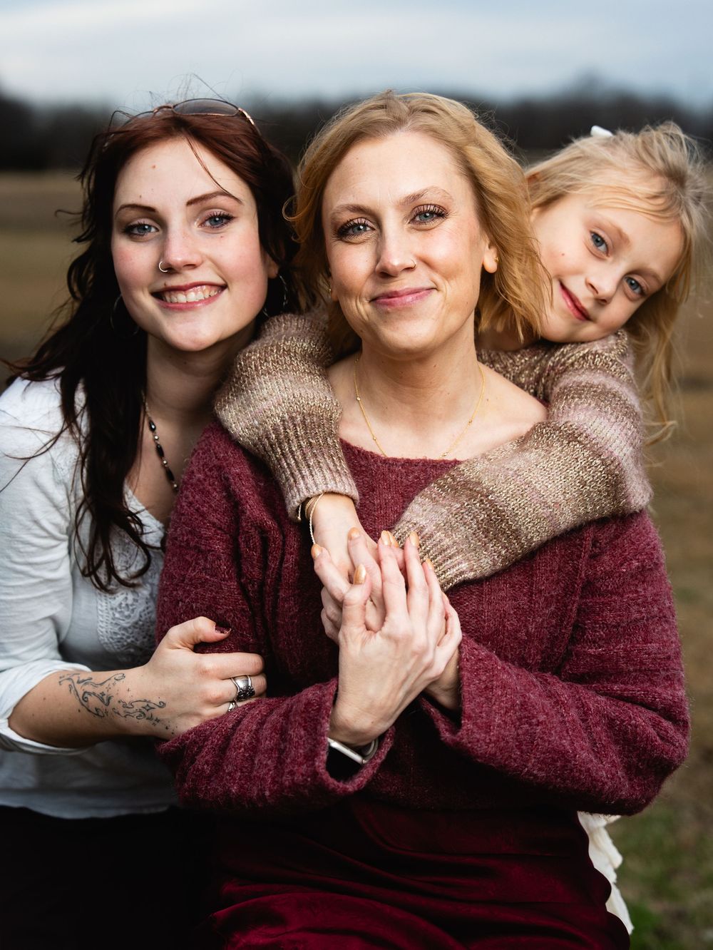 A woman in a burgundy sweater poses outdoors with two daughters, one teenage and one young child, in a warm family portrait.