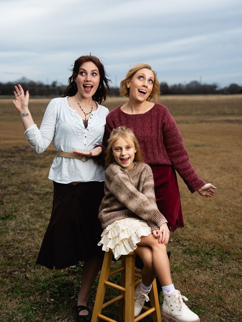 Two women and a young girl pose playfully in an open field, wearing cozy sweaters and skirts for a fun outdoor photo.