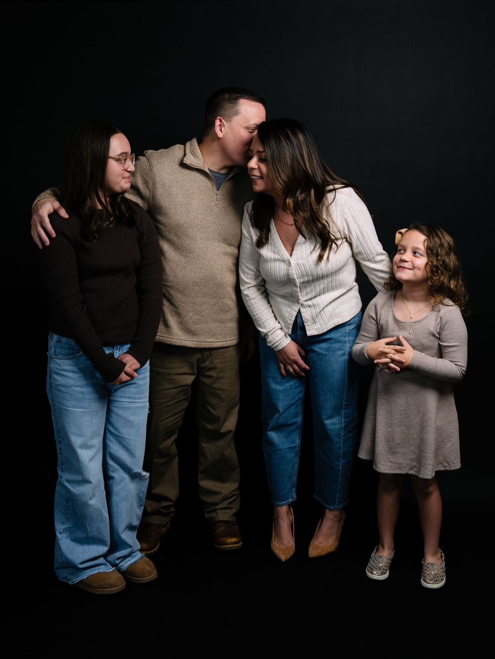 A happy family of four poses together against a dark background in a warm studio portrait session.