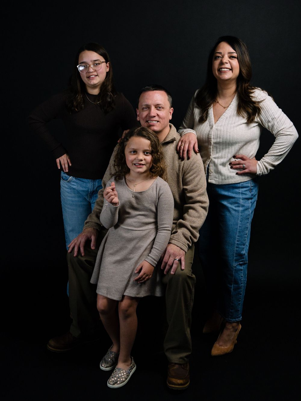 Family of four posing together against dark background, parents with two daughters smiling in casual attire.