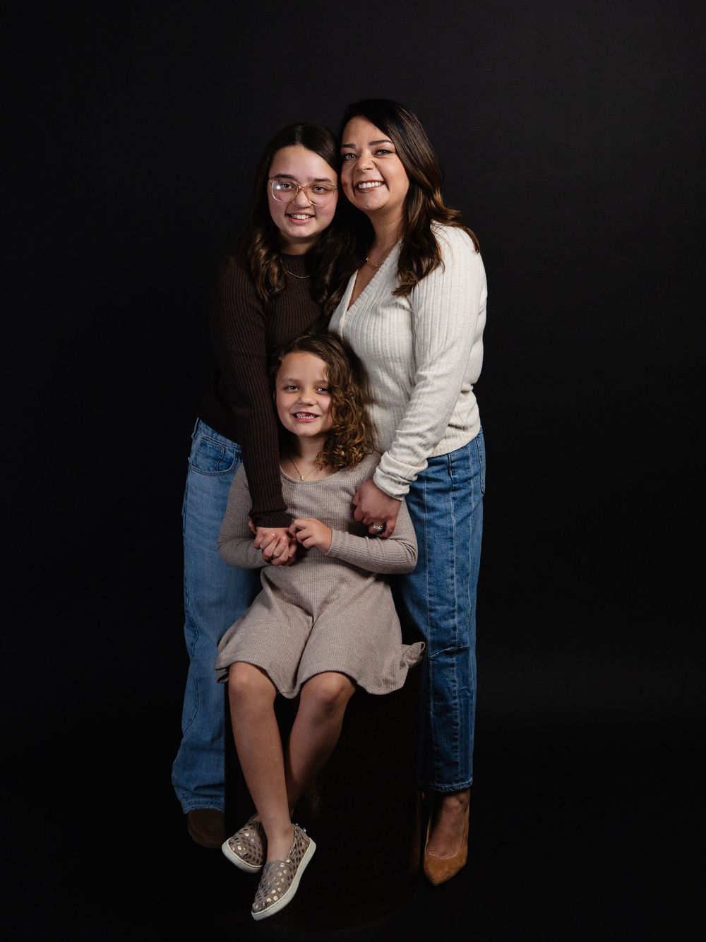 Studio portrait of three smiling females, two teens and a young girl, posing together against a dark background.